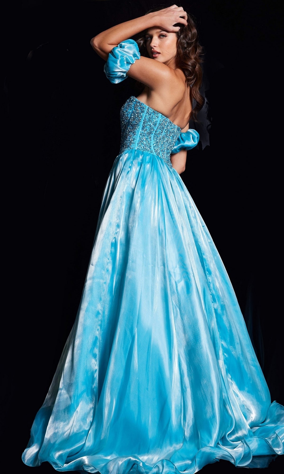 A woman poses against a black background in the Long Prom Dress 36591 by Jovani, a shiny light blue floor-length A-line gown with a fitted beaded corset bodice and full skirt.